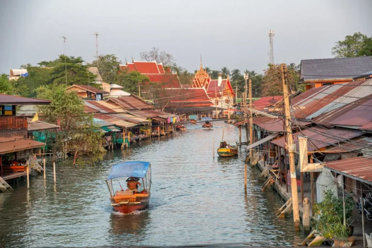 Bangkok et le Centre 16 Canal d'Amphawa bordé de maisons en bois sur pilotis, avec un bateau longtail au premier plan et un temple aux toits rouges en arrière-plan, près de Bangkok