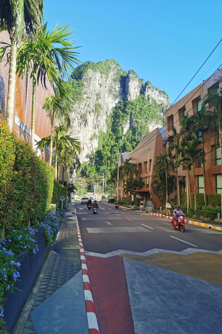 Rue animée d'Ao Nang bordée de palmiers avec une imposante falaise calcaire en arrière-plan sous un ciel bleu dégagé.