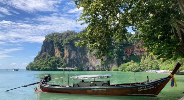 Bateau traditionnel à longue queue amarré à Railay, avec d'imposantes falaises calcaires couvertes de jungle en arrière-plan et une eau vert émeraude, Thaïlande.