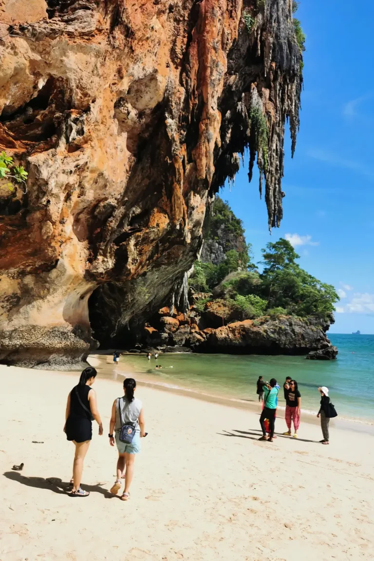TouristEs marchant sur Phra Nang Beach à Railay, face à une imposante falaise calcaire et une grotte, Thaïlande.