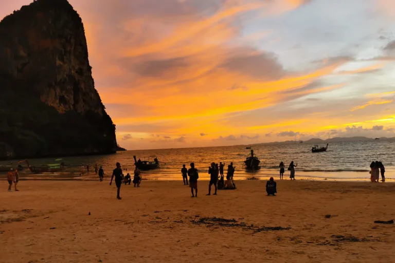 Silhouette de touristes et de bateaux long-tail sur la plage de Railay West pendant un spectaculaire coucher de soleil orange et doré.