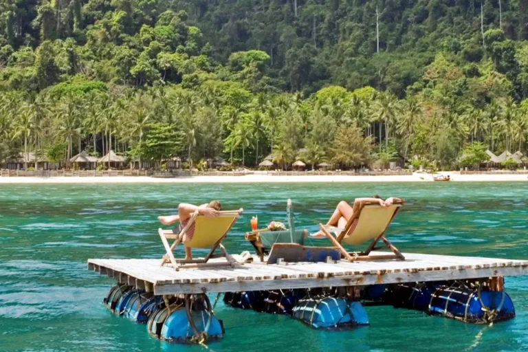 Essence du Sud 11 Couple se relaxant sur un ponton flottant en mer turquoise face à l'éco-resort Coco Cottage sur l'île de Koh Ngai, avec plage et cocotiers en arrière-plan